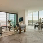 Property for sale Modern, bright apartment in Langford Tower Sheffield with large windows offering a city view, a cozy living area with sofas and a rug, a dining table with chairs, and neutral, stylish décor throughout. Presented by Palace Auctions