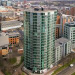 Property for sale A tall, modern glass apartment building with rounded edges stands at an intersection in the urban cityscape—ideal for anyone seeking new build homes in Sheffield or a prime investment property. Roads and cars are visible below amidst other city buildings. Presented by Palace Auctions