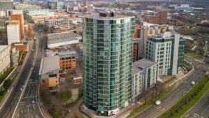 Property for sale A tall, modern glass apartment building with rounded edges stands at an intersection in the urban cityscape—ideal for anyone seeking new build homes in Sheffield or a prime investment property. Roads and cars are visible below amidst other city buildings. Presented by Palace Auctions