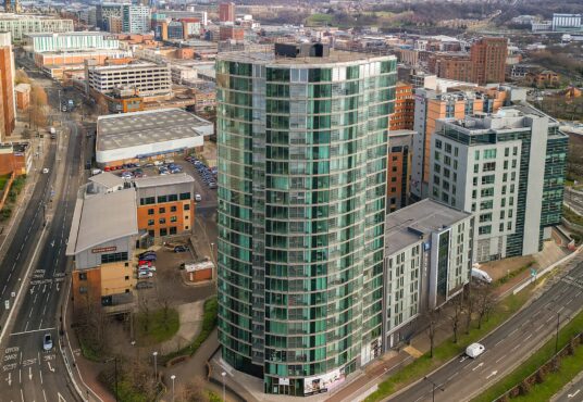Property for sale A tall, modern glass apartment building with rounded edges stands at an intersection in the urban cityscape—ideal for anyone seeking new build homes in Sheffield or a prime investment property. Roads and cars are visible below amidst other city buildings. Presented by Palace Auctions