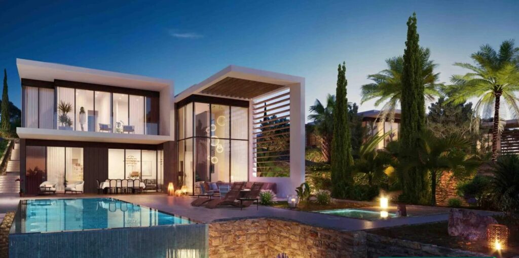 Property for sale Modern two-story house with large windows, a swimming pool, lounge chairs, and lush green trees—an elegant example of Pegeia luxury real estate, beautifully illuminated by outdoor lighting at dusk. Presented by Palace Auctions