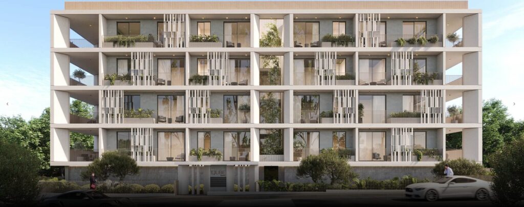 Property for sale A modern four-story mixed-use apartment building in Paphos Centre, featuring large balconies, glass railings, and lush greenery. Trees and plants enhance the exterior, while cars and a pedestrian can be seen in front of the building. Presented by Palace Auctions