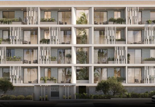 Property for sale A modern four-story mixed-use apartment building in Paphos Centre, featuring large balconies, glass railings, and lush greenery. Trees and plants enhance the exterior, while cars and a pedestrian can be seen in front of the building. Presented by Palace Auctions