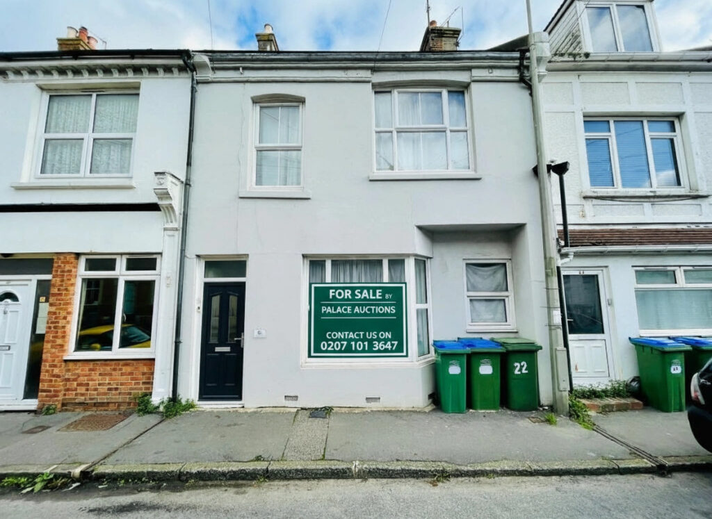 Property for sale A two-story terraced house with a black front door and large "For Sale" sign in the window—ideal as a 2-bed maisonette Newhaven investment. Three green recycling bins stand out front, with neighboring houses attached on both sides. Presented by Palace Auctions