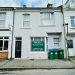 Property for sale A two-story terraced house with a black front door and large "For Sale" sign in the window—ideal as a 2-bed maisonette Newhaven investment. Three green recycling bins stand out front, with neighboring houses attached on both sides. Presented by Palace Auctions