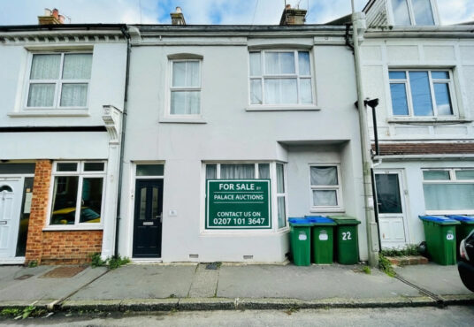Property for sale A two-story terraced house with a black front door and large "For Sale" sign in the window—ideal as a 2-bed maisonette Newhaven investment. Three green recycling bins stand out front, with neighboring houses attached on both sides. Presented by Palace Auctions