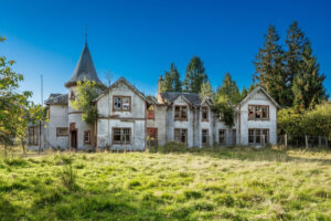 Property for sale An abandoned, overgrown mansion with boarded windows, peeling paint, and a small turret sits in a grassy field under a bright blue sky, surrounded by tall trees—a unique development opportunity near Loch Lomond. Presented by Palace Auctions