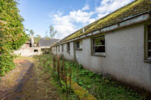 Property for sale An abandoned, long, single-story building with peeling paint and moss-covered roof, overgrown plants, broken windows, and a cracked pathway—once part of a Scottish manor house estate now offering potential for income-producing development. Presented by Palace Auctions