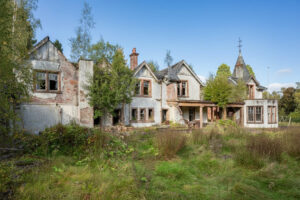 Property for sale An old, abandoned two-story house with broken windows and crumbling walls sits surrounded by overgrown grass, evoking a forgotten Scottish manor house development under a clear blue sky. Presented by Palace Auctions