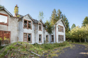 Property for sale An abandoned, two-story house with broken windows and a damaged roof sits among overgrown vegetation, offering a unique development opportunity near Loch Lomond beneath a clear blue sky. Presented by Palace Auctions