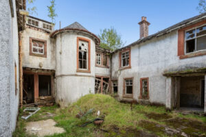 Property for sale A dilapidated, abandoned two-story house—once a grand Scottish manor house for sale—with broken windows, overgrown grass, and crumbling walls surrounds a small courtyard under a clear blue sky. Presented by Palace Auctions