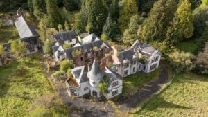 Property for sale Aerial view of a large, abandoned Scottish manor house development with turrets and overgrown vegetation; the building’s roof is damaged and surrounding grounds, bordered by tall trees, hint at a unique development opportunity Loch Lomond. Presented by Palace Auctions