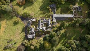 Property for sale Aerial view of an old, abandoned stone building complex—once a grand Scottish manor house development—surrounded by trees and grassy fields, with collapsed roofs and overgrown vegetation encroaching on the historic structure. Presented by Palace Auctions