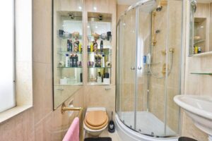 Property for sale A compact bathroom in an Albion Gate apartment with a glass shower, tan tile walls, a toilet with wooden seat, a white sink, and mirrored shelves holding toiletries—perfect for those seeking a two bedroom Hyde Park residence. A pink towel hangs by the window. Presented by Palace Auctions