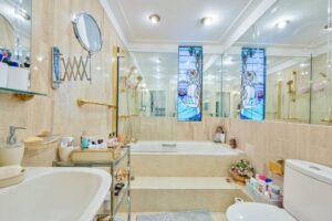 Property for sale A bright bathroom in this Central London flat W2 features beige tiles, a bathtub with steps, wall mirrors, gold fixtures, and a stained-glass window. Toiletries and decor are neatly arranged on shelves and a rolling cart. Presented by Palace Auctions