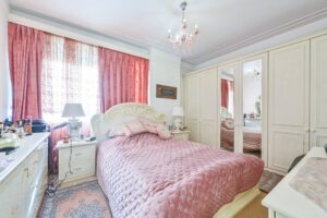 Property for sale A bright, elegant bedroom in an Albion Gate apartment features a large bed with a pink quilt, matching pink curtains, a chandelier, mirrored wardrobes, white furniture, and decorative pillows. Presented by Palace Auctions