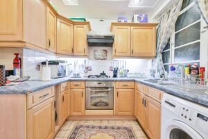 Property for sale A tidy, well-lit kitchen with light wooden cabinets, gray countertops, gas stove, oven, washing machine, and large window with patterned curtains—an inviting feature of this Central London flat W2 near Hyde Park apartment auction. Presented by Palace Auctions