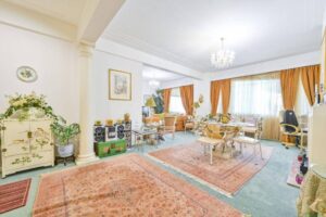 Property for sale A spacious, elegant living and dining room in a Central London flat W2, with large windows, gold curtains, ornate chandeliers, vintage furniture, patterned rugs, and decorative plants. The room has a bright, classic ambiance. Presented by Palace Auctions