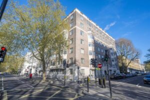 Property for sale A large, light-brown brick apartment building stands on a street corner under a clear blue sky, with trees lining the sidewalk and traffic lights at the intersection—an ideal Central London flat W2 near the upcoming Bayswater property auction. Presented by Palace Auctions