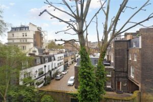Property for sale View of a quiet, cobblestone residential street lined with white and brown brick townhouses near a sought-after Two bedroom Hyde Park, leafy trees, and a partially leafless tree in the foreground under a blue sky with scattered clouds. Presented by Palace Auctions