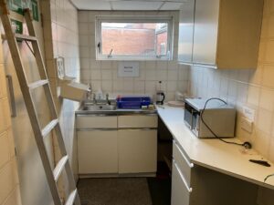 Property for sale A small kitchen with white tiled walls, a sink under a window, dish drainer with blue tray, microwave, and kettle. A ladder leans against the wall on the left. Ideal for an Eastwood investment property or Mansfield Road property. "CAUTION" sign on window. Presented by Palace Auctions