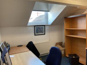 Property for sale Small office room with slanted ceiling, two wooden desks, blue chairs, empty shelves, computer monitor, cardboard boxes, framed picture, and a window with blinds—ideal as an Eastwood investment property or listed in a Nottinghamshire auction. Presented by Palace Auctions