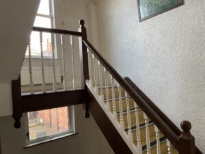 Property for sale A wooden staircase with white balusters and a dark brown handrail leads up to a landing with a window letting in natural light—an inviting detail of this Mansfield Road property, ideal as a development or investment property. A textured white wall and framed painting are visible. Presented by Palace Auctions