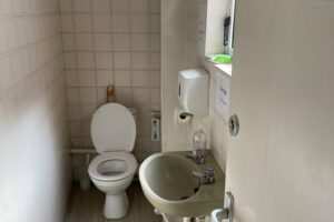 Property for sale A small bathroom in a Mansfield Road property features a white toilet with an open lid, beige basin, soap dispenser, and hand sanitiser bottle. The tiled wall and window above the basin add charm to this potential development or investment property. Presented by Palace Auctions