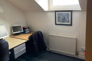Property for sale A small office with a desk, computer, stacks of files, and a black chair with a jacket draped over it—ideal for those seeking an Eastwood investment property on Mansfield Road—with a radiator, window, and framed picture on a white wall. Presented by Palace Auctions