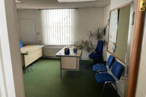Property for sale Small office with green carpet, two desks, blue chairs, large window with vertical blinds, whiteboard, noticeboard, and potted plants in the corner. Sunlight enters through the window—ideal for an Eastwood investment property. Presented by Palace Auctions