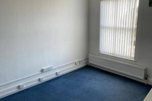 Property for sale A small, empty room with white walls, a blue carpet, large window with vertical blinds, radiator below the window, and multiple sockets—ideal as a development or investment property in an upcoming Nottinghamshire auction. Presented by Palace Auctions