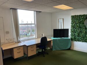 Property for sale Bright office with two wooden desks, a blue office chair, a window with white curtains, TV on a green-clothed table, wall mirror, and a vertical panel of artificial greenery—ideal for an Eastwood investment property or Nottinghamshire auction listing. Presented by Palace Auctions