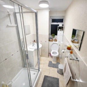 Property for sale Bright, modern bathroom in a one-bedroom flat Nottingham with a glass shower, white sink, toilet, shelves with plants and toiletries, large mirror, towel rack, and gray floor mats on beige tiles. Presented by Palace Auctions
