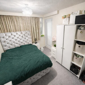 Property for sale A tidy one bedroom flat Nottingham with a large bed featuring a tufted silver headboard and dark green cover, white wardrobes, shelves with storage baskets, beige curtains, gray carpet, and a small chandelier light fixture. Presented by Palace Auctions