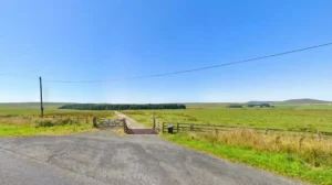 Property for sale A paved road leads to a wooden gate opening onto a narrow path through green fields under a clear blue sky, with distant trees and low hills—an inviting land for sale near Bolventor, Bodmin, Cornwall. Presented by Palace Auctions