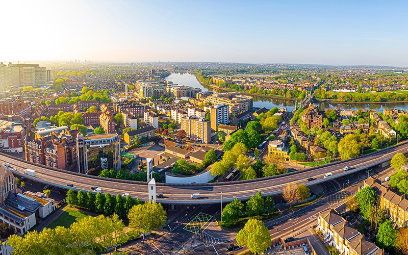 Property for sale A panoramic aerial view of a city, featuring modern and older buildings, green trees, a river in the background, and a main road curving through the foreground—showcasing the vibrant area known for Hammersmith and Fulham property auctions on a clear, sunny day. Presented by Palace Auctions
