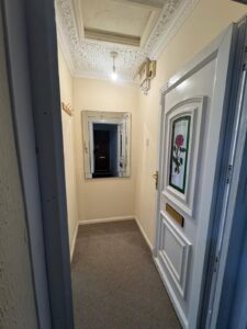 Property for sale Narrow hallway in Wynyard Mews with an ornate white ceiling, hanging lightbulb, decorative white door with floral panel, wall mirror, beige walls, grey carpet, and a small coat rack—ideal for a buy-to-let investment amid Hartlepool regeneration. Presented by Palace Auctions