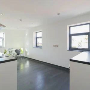 Property for sale A modern, minimalistic kitchen with white walls, dark flooring, black worktops, and two windows—ideal for Coventry PBSA Investment Auction listings. In the corner, a small dining table seats four; some chairs feature striking green backs. Presented by Palace Auctions