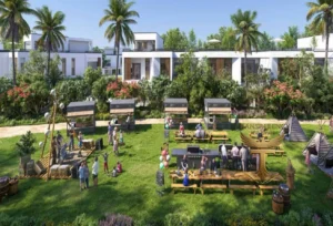 Property for sale A vibrant outdoor scene at DAMAC Riverside shows people gathered on a green lawn with picnic tables, food stalls, balloons, and children playing, surrounded by palm trees and modern white buildings near Dubai Investment Park. Presented by Palace Auctions