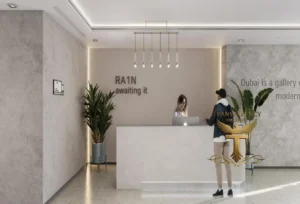 Property for sale A modern reception area at Ra1n Residence JVC flat features a white desk, two potted plants, a woman working at the desk, and a man standing in front. "RA1N awaiting it" is on the wall, with contemporary lighting above. Presented by Palace Auctions