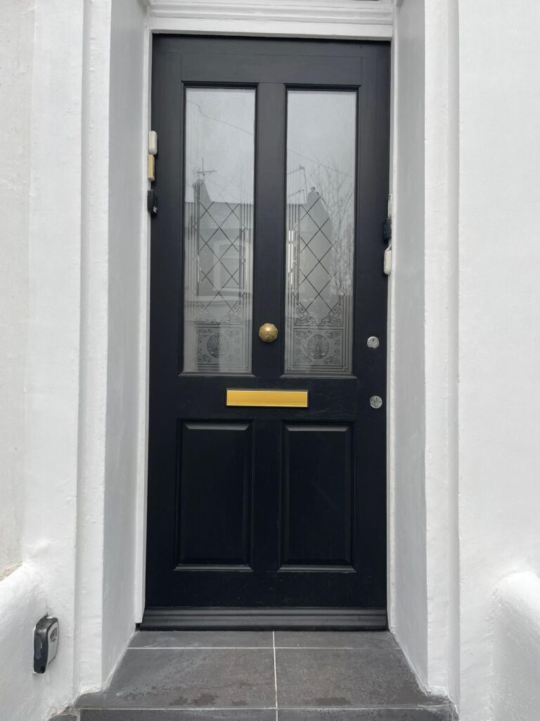Property for sale A black wooden door with frosted glass panels featuring diamond patterns and decorative motifs, a brass letterbox, round door knob, and doorbell, set in a white painted doorway—an inviting entrance to a Poets' Corner ground floor flat. Presented by Palace Auctions