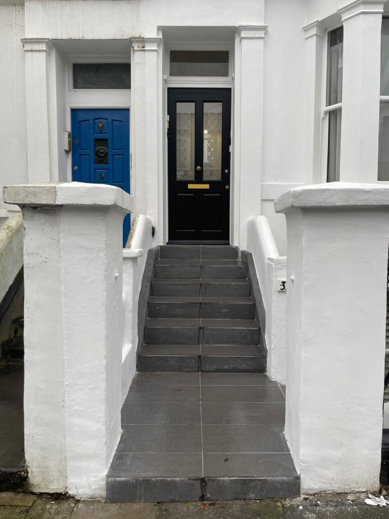 Property for sale A small set of grey steps leads to a black front door with glass panels and a gold letterbox. The entrance to this Poets' Corner ground floor flat is framed by white walls, with a blue door visible to the left. Presented by Palace Auctions