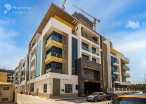 Property for sale Modern multi-storey Ra1n Residence JVC apartment building with yellow and white accents, glass balconies, and a large entrance. Several cars are parked in front, with cranes on the rooftop against a blue sky. Property Finder logo in the corner. Presented by Palace Auctions