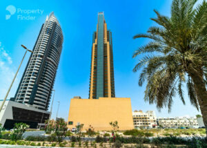 Property for sale A tall modern skyscraper with a glass facade stands between a curved high-rise and a palm tree, against a clear blue sky—an impressive view ideal for a Dubai investment flat 2026. Smaller buildings and greenery complete this luxury cityscape. Presented by Palace Auctions