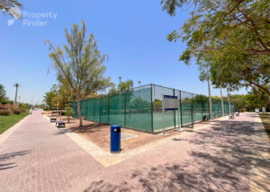 Property for sale A sunny outdoor tennis court in Jumeirah Village Triangle, surrounded by green fencing, trees, benches, and a walking path. The clear blue sky adds to the peaceful ambience of this well-maintained luxury flat community. Presented by Palace Auctions