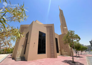 Property for sale A beige mosque with tall, narrow windows and a modern minaret stands under a clear blue sky in Jumeirah Village Triangle, surrounded by small trees and set on a pink-tiled walkway near luxury flats Dubai is known for. Presented by Palace Auctions