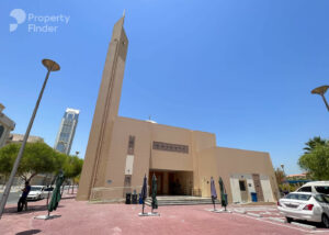 Property for sale A beige mosque with a tall minaret stands under a clear blue sky in Jumeirah Village Triangle, Dubai, surrounded by trees, street lamps, parked cars, and a pink-tiled courtyard. The Property Finder logo appears in the top left corner. Presented by Palace Auctions