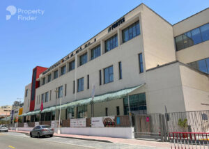 Property for sale A modern three-storey building with large windows and a shaded entrance in Jumeirah Village Triangle, featuring English and Arabic signage. Cars line the street beneath a clear blue sky, highlighting this luxury flat in Dubai. Presented by Palace Auctions