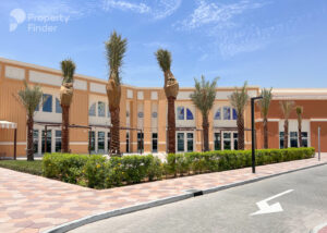 Property for sale A modern, orange-and-cream Capri 2 building with tall palm trees, manicured bushes, and a paved walkway under a blue sky. The scene is sunny and bright, offering DAMAC Riverside Views through the building’s large windows. Presented by Palace Auctions