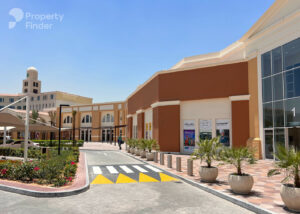 Property for sale A modern commercial building with glass entrances, potted plants, and clear blue sky, featuring shopfronts and a drive with yellow speed bumps. The DAMAC Riverside Views logo is visible alongside "Property Finder" in the top left corner. Presented by Palace Auctions